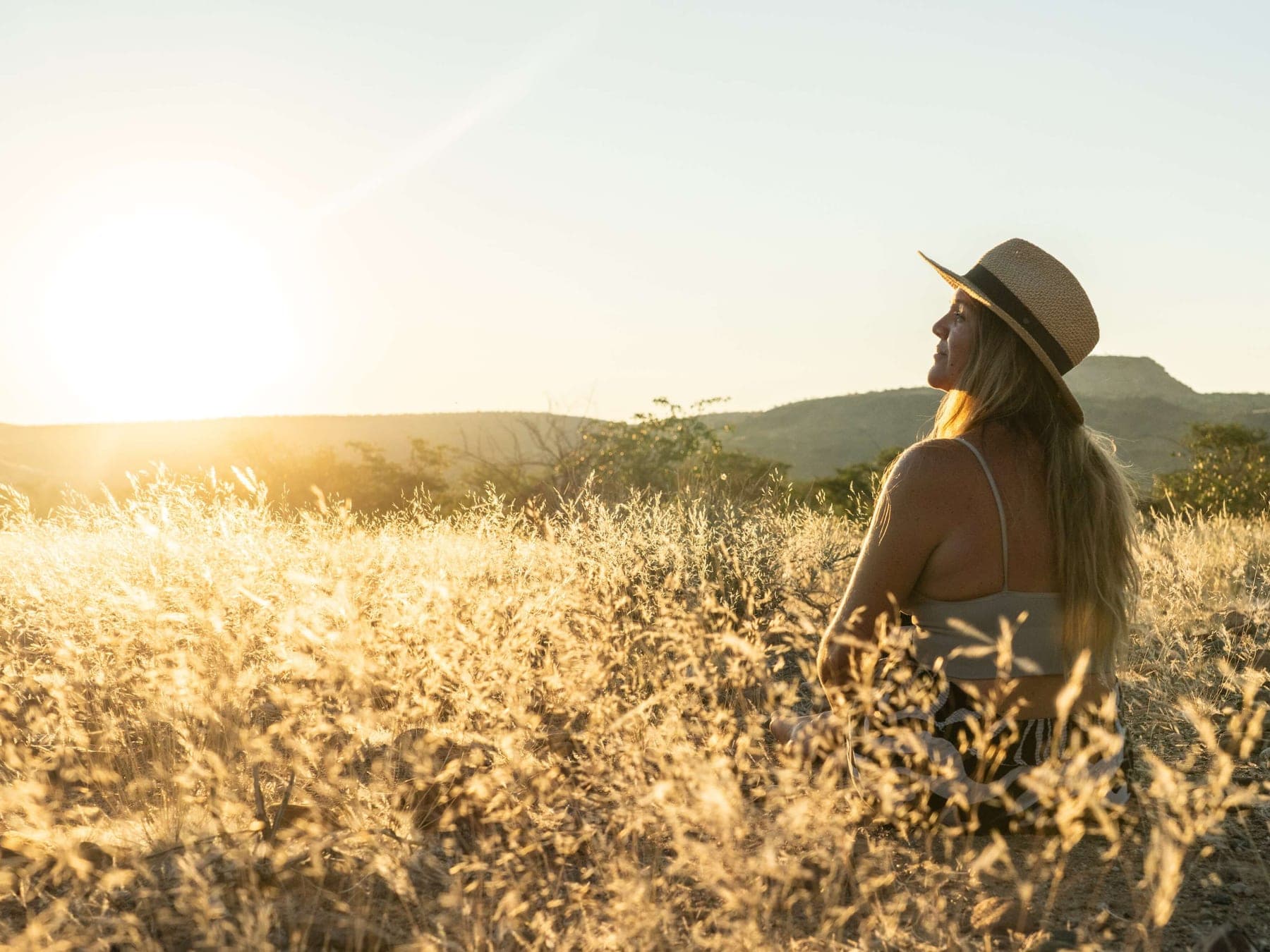 Léa, chapeau de paille, assise dans les herbes dorées face au soleil